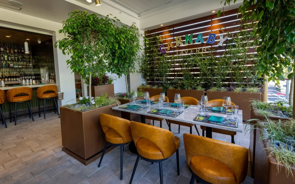 Garden terrace dining area at Gordon Ramsay at The Carnaby, featuring lush greenery, tan seating, a set dining table, and Carnaby Tavern signage.