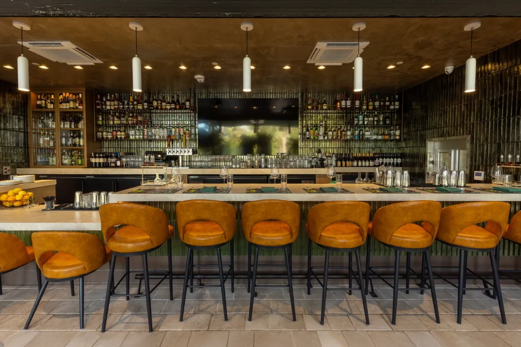 Bar at Gordon Ramsay at The Carnaby with tan barstools, bottle-lined shelves, pendant lighting, and a long stone counter.