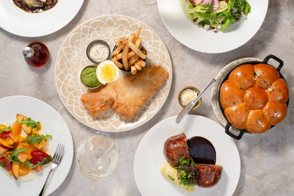 Overhead spread of Gordon Ramsay At The Carnaby dishes, including fish and chips, salad, bread rolls, roasted meat and seasonal vegetables.